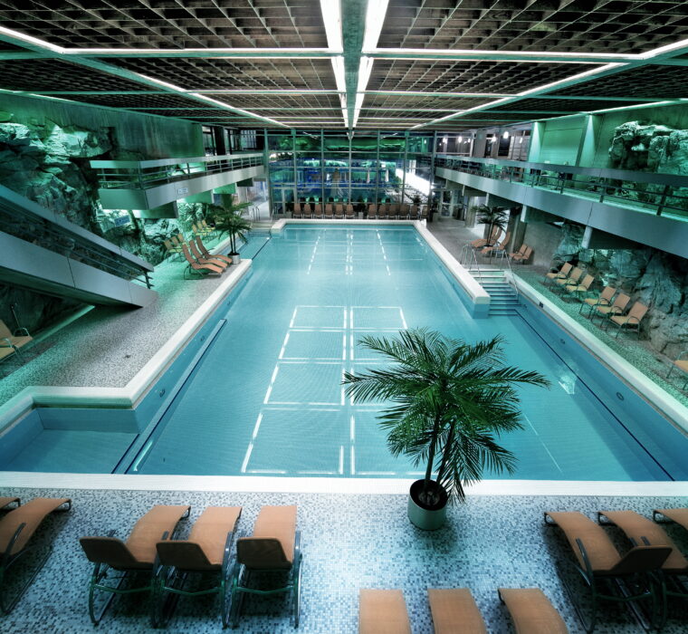 Innenaufnahme eines großen Schwimmbeckens in der Felsentherme Gastein mit Liegestühlen und Felswänden für Erholung und