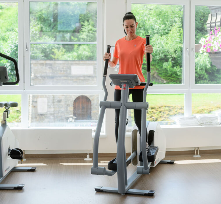Frau nutzt im modernen Fitnessraum des Bärenhofs einen Crosstrainer für gelenkschonendes Ausdauertraining, im Hintergrund Blick nach draußen auf Garten und Steinmauer.