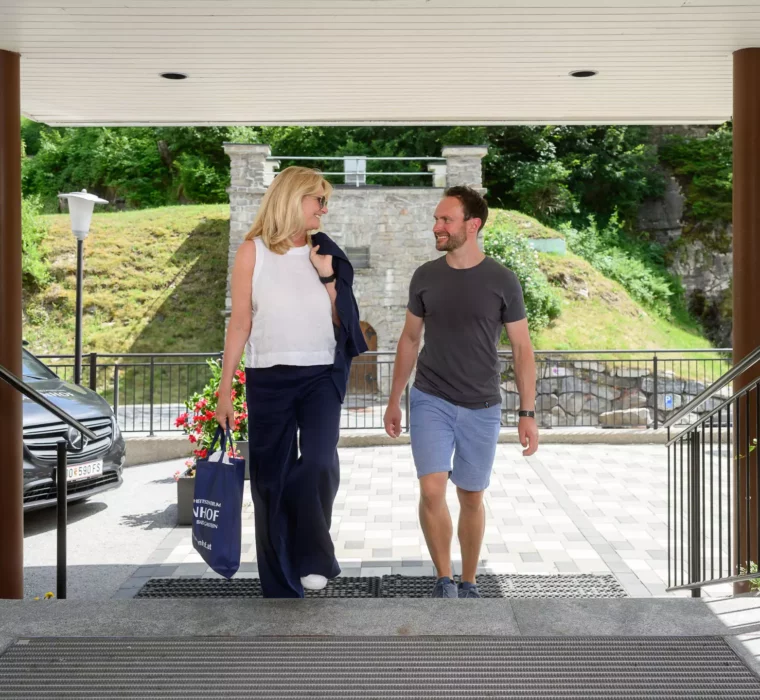 Zwei Menschen betreten gemeinsam das Gesundheitszentrum Bärenhof in Bad Gastein – freundlicher Empfangsbereich mit barrierefreiem Zugang und Naturkulisse im Hintergrund.