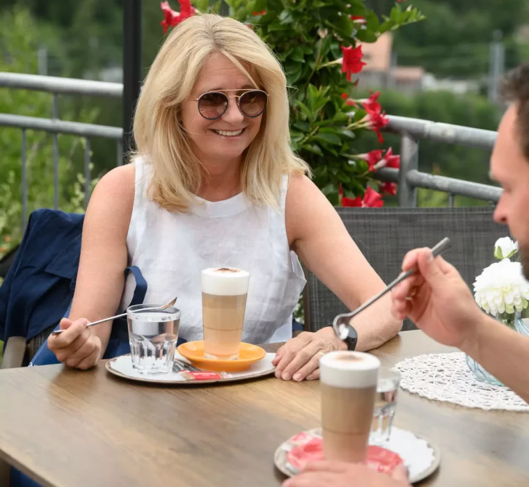 Frau genießt mit Begleitung einen Latte Macchiato auf der Panoramaterrasse im Felsencafé des Bärenhofs in Bad Gastein – entspannte Atmosphäre mit Blick ins Grüne.
