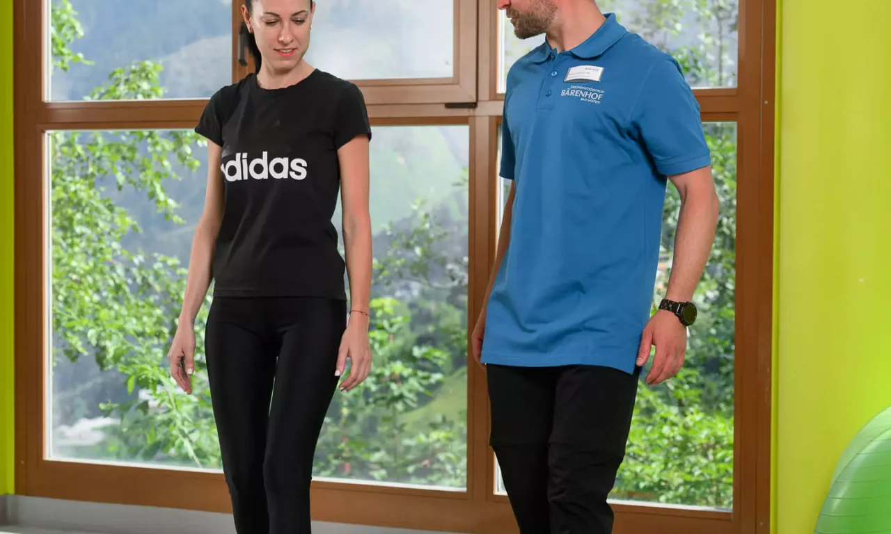 Physiotherapeut zeigt Patientin eine Übung mit Theraband auf Gymnastikmatte vor Panoramafenster mit Bergblick.