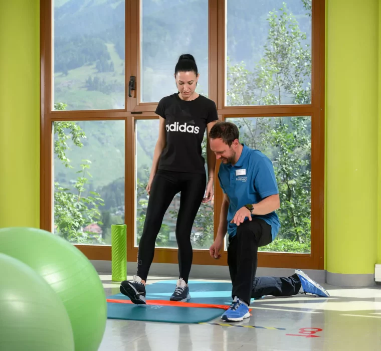 Physiotherapeut zeigt Patientin eine Übung mit Theraband auf Gymnastikmatte vor Panoramafenster mit Bergblick.