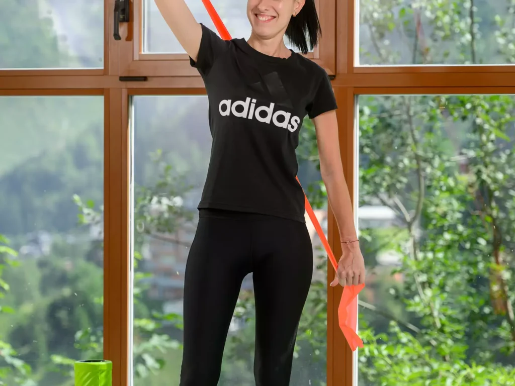 Patientin führt eine therapeutische Schulterübung mit Theraband in heller Trainingsumgebung mit Naturblick durch.