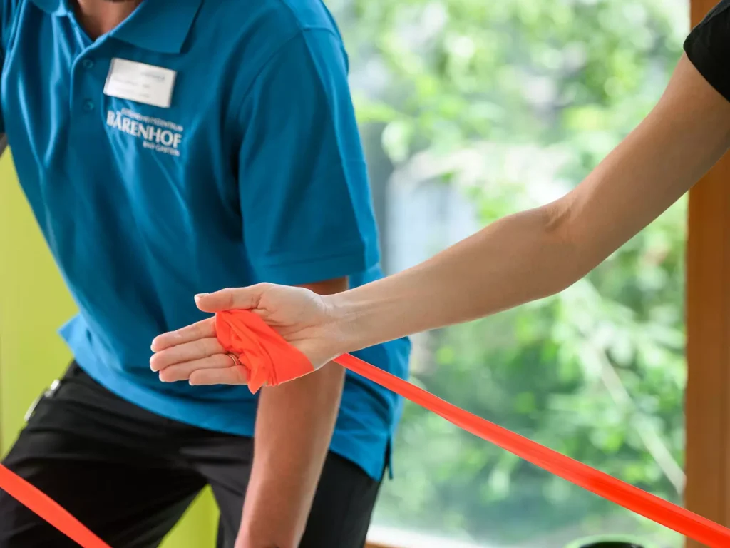 Physiotherapeut zeigt Patientin eine Übung mit Theraband auf Gymnastikmatte vor Panoramafenster mit Bergblick.