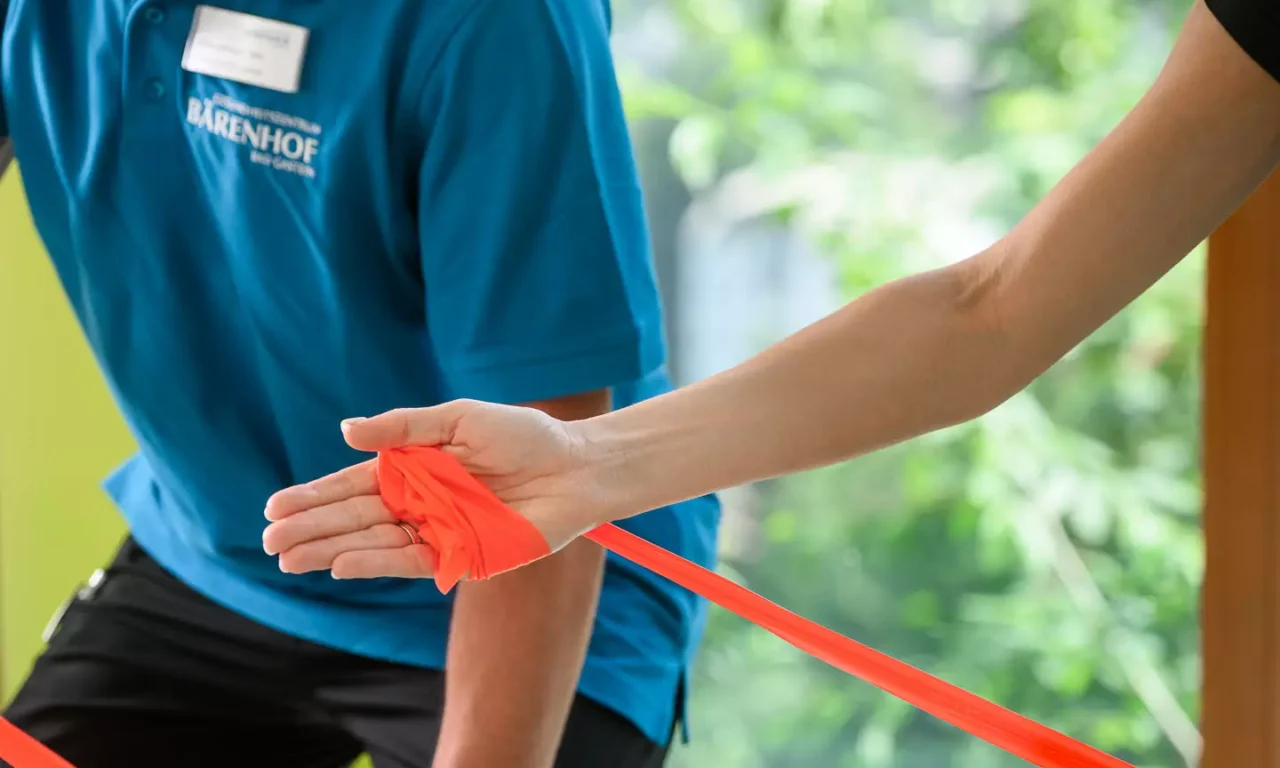 Physiotherapeut zeigt Patientin eine Übung mit Theraband auf Gymnastikmatte vor Panoramafenster mit Bergblick.