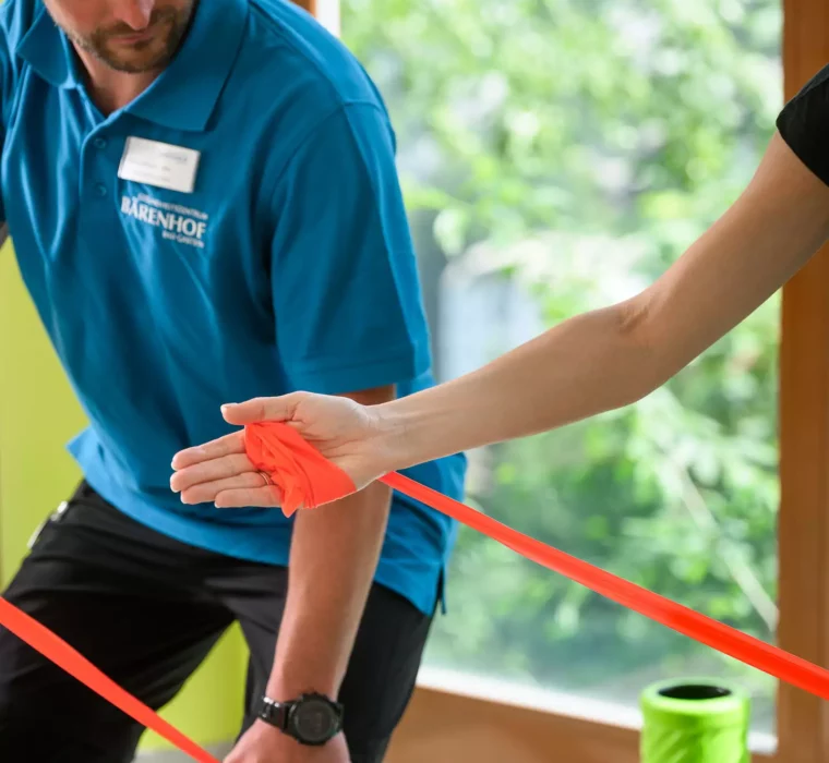 Physiotherapeut zeigt Patientin eine Übung mit Theraband auf Gymnastikmatte vor Panoramafenster mit Bergblick.
