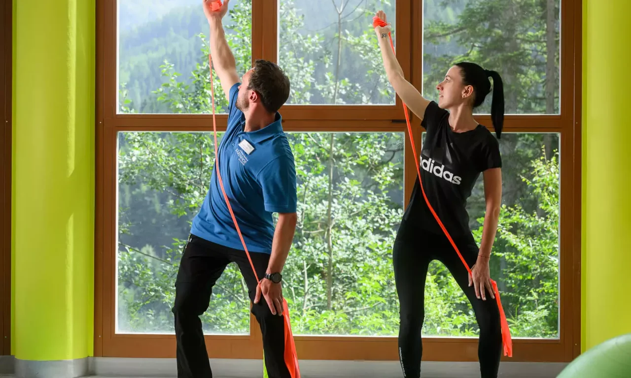 Physiotherapeut zeigt Patientin eine Übung mit Theraband auf Gymnastikmatte vor Panoramafenster mit Bergblick.