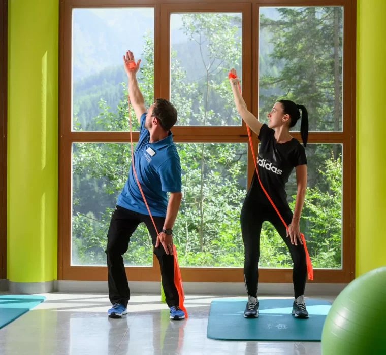 Physiotherapeut zeigt Patientin eine Übung mit Theraband auf Gymnastikmatte vor Panoramafenster mit Bergblick.