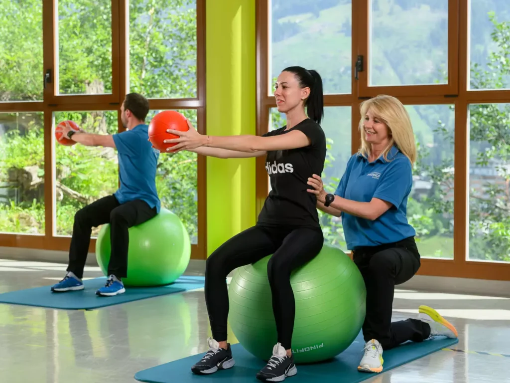 Therapeutin begleitet Patientin bei Gleichgewichts- und Rumpfstabilitätsübung auf dem Gymnastikball mit Pezziball und Ball im hellen Therapieraum mit Ausblick auf die Berge.