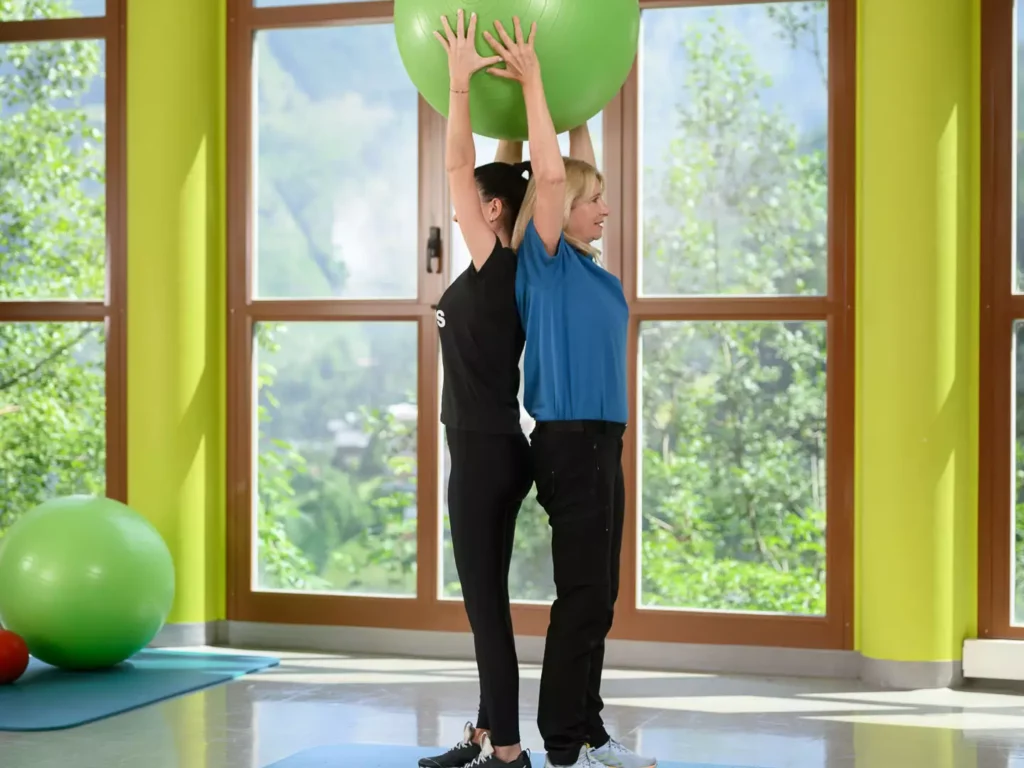 Therapeutin und Patientin führen Rücken-an-Rücken-Übung mit Gymnastikball zur Förderung von Koordination und Körperspannung im sonnigen Trainingsraum mit Bergblick durch.