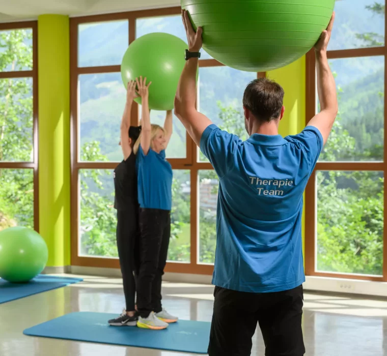 Therapie-Team begleitet Patientinnen bei gemeinsamer Übung mit Gymnastikbällen zur Förderung von Beweglichkeit und Haltung im lichtdurchfluteten Trainingsraum mit Bergpanorama.