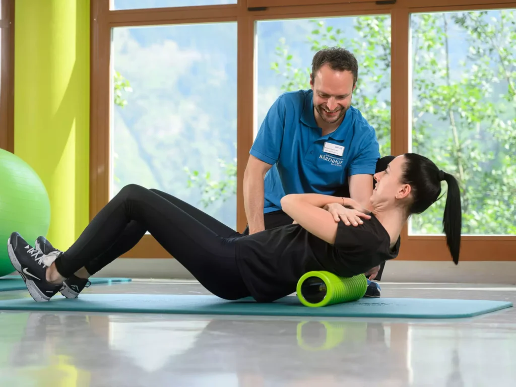 Therapeut vom Bärenhof unterstützt Patientin einfühlsam bei Rückenübung mit Faszienrolle im hellen Trainingsraum mit Naturausblick.