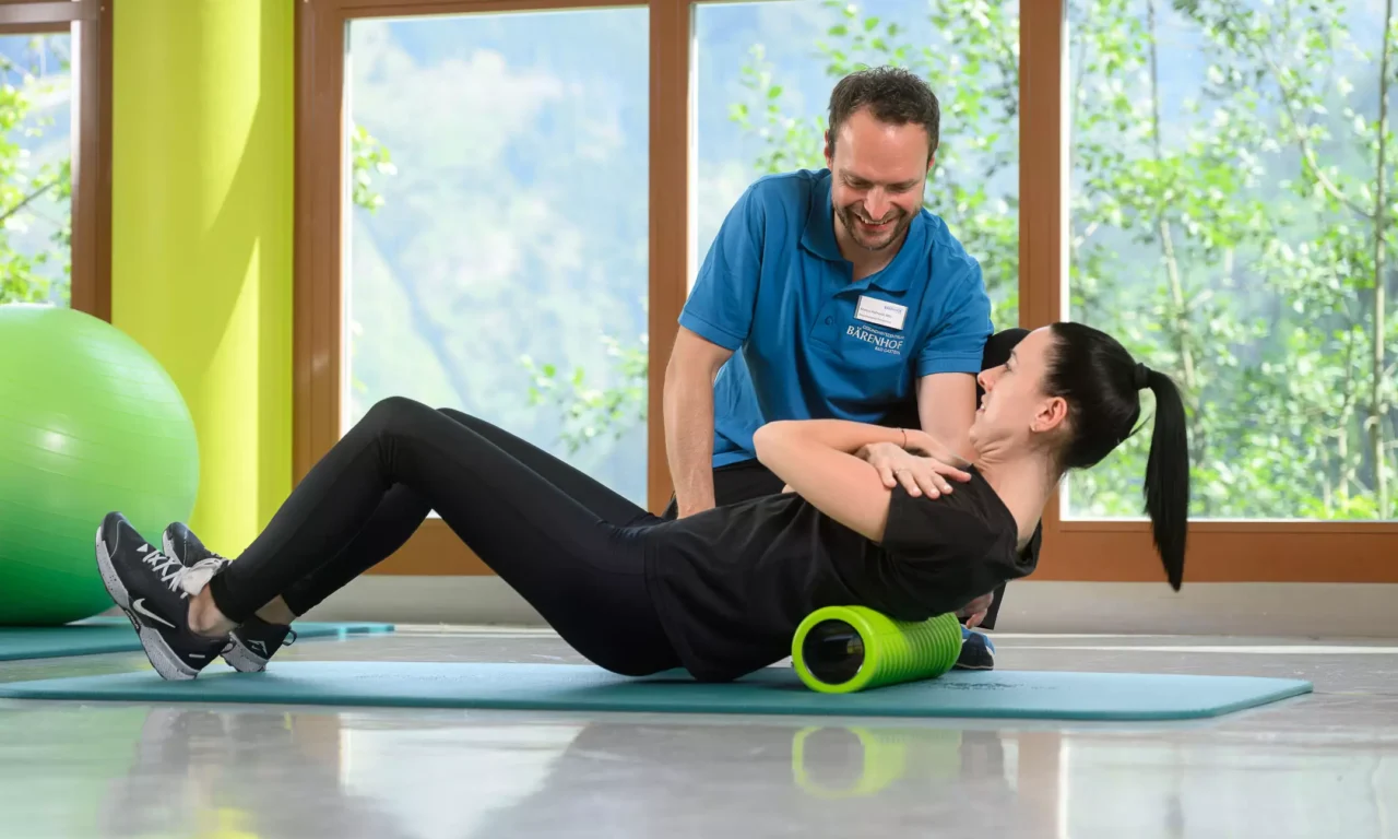 Therapeut vom Bärenhof unterstützt Patientin einfühlsam bei Rückenübung mit Faszienrolle im hellen Trainingsraum mit Naturausblick.