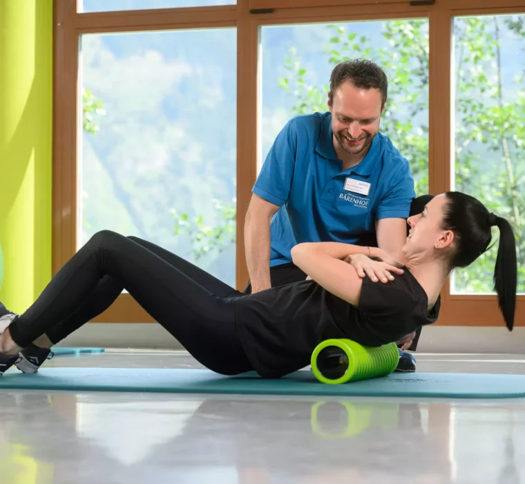 Therapeut vom Bärenhof unterstützt Patientin einfühlsam bei Rückenübung mit Faszienrolle im hellen Trainingsraum mit Naturausblick.