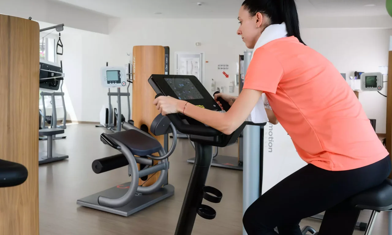 Patientin trainiert im modernen Fitnessbereich des Bärenhofs auf einem Technogym-Ergometer mit digitalem Display.