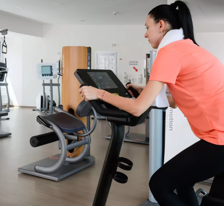 Patientin trainiert im modernen Fitnessbereich des Bärenhofs auf einem Technogym-Ergometer mit digitalem Display.