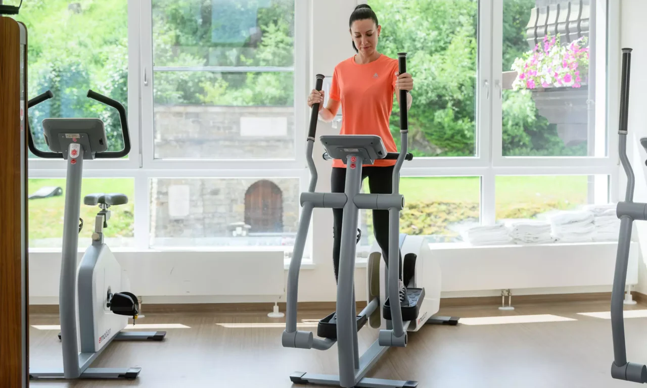Frau nutzt im modernen Fitnessraum des Bärenhofs einen Crosstrainer für gelenkschonendes Ausdauertraining, im Hintergrund Blick nach draußen auf Garten und Steinmauer.