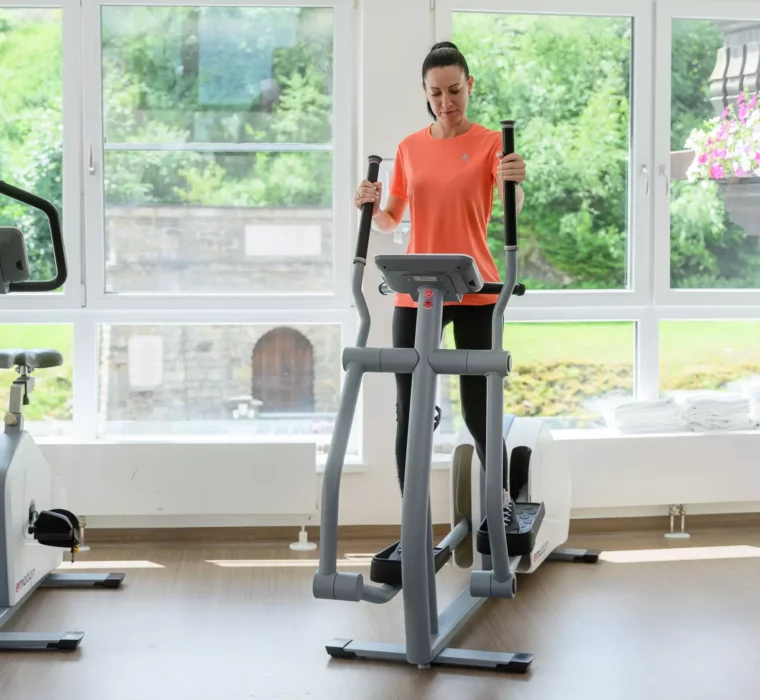 Frau nutzt im modernen Fitnessraum des Bärenhofs einen Crosstrainer für gelenkschonendes Ausdauertraining, im Hintergrund Blick nach draußen auf Garten und Steinmauer.