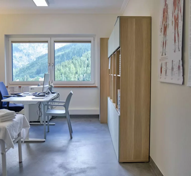Behandlungszimmer im Gesundheitszentrum Bärenhof Bad Gastein mit Therapie- und Untersuchungsliege, Schreibtisch und Bergblick