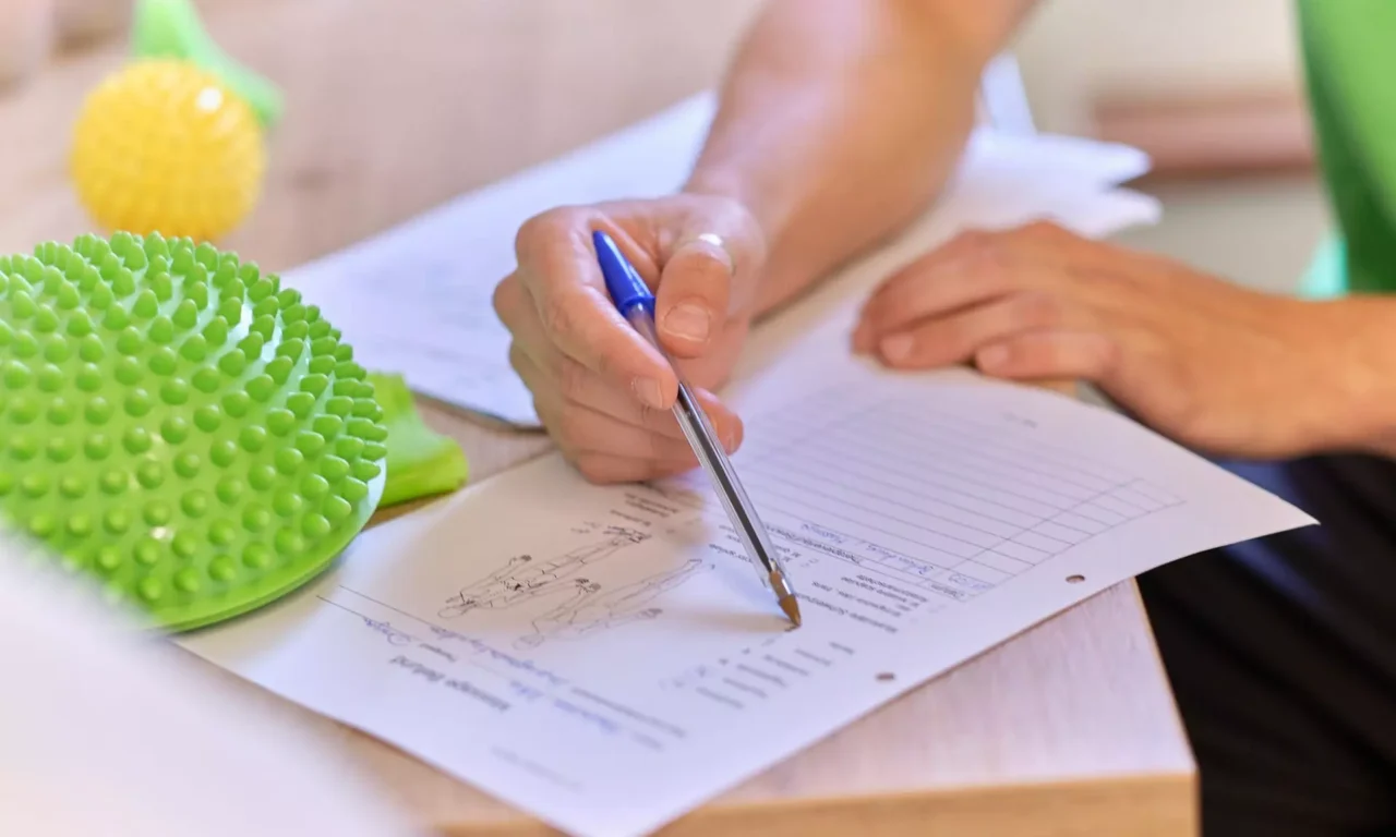 Hand mit Kugelschreiber zeigt auf ein Formular mit anatomischen Zeichnungen auf einem Holztisch im Gesundheitszentrum