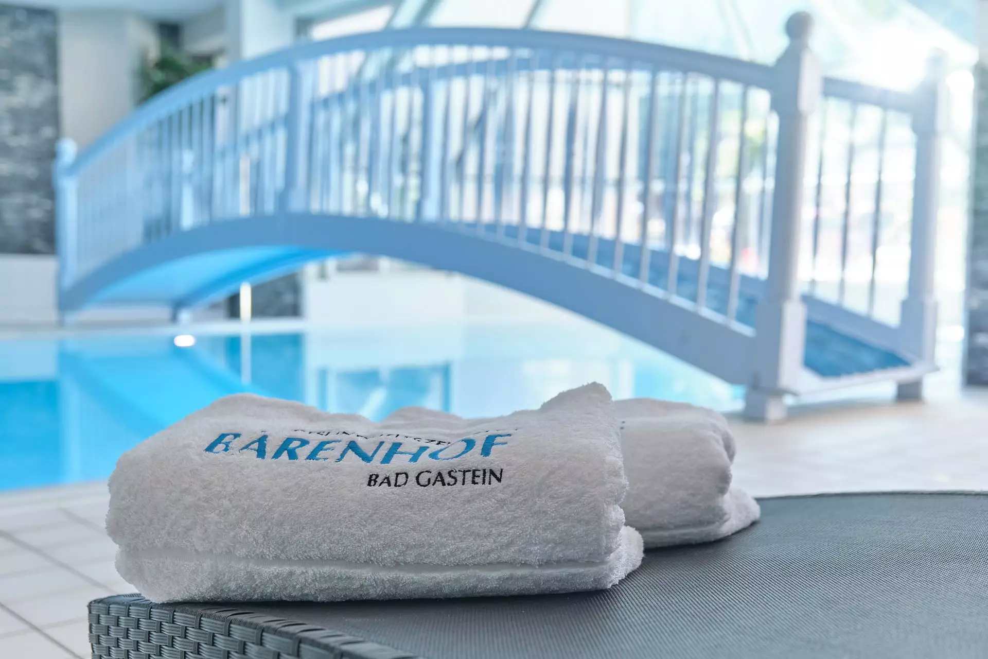 Weißes Handtuch mit Aufschrift Bärenhof Bad Gastein am Rand eines Hallenbads mit blauer Brücke im Hintergrund