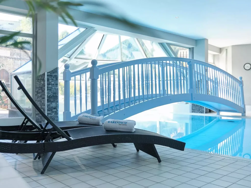 Innenaufnahme des Hallenbads im Gesundheitszentrum Kurhotel Bärenhof Bad Gastein mit Liegen und weißer Brücke über dem Pool