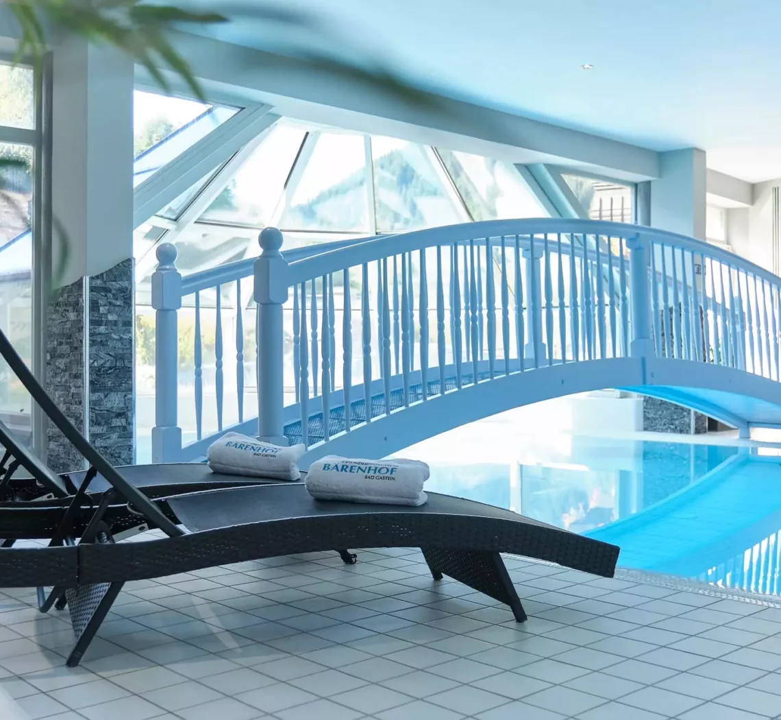 Innenaufnahme des Hallenbads im Gesundheitszentrum Kurhotel Bärenhof Bad Gastein mit Liegen und weißer Brücke über dem Pool