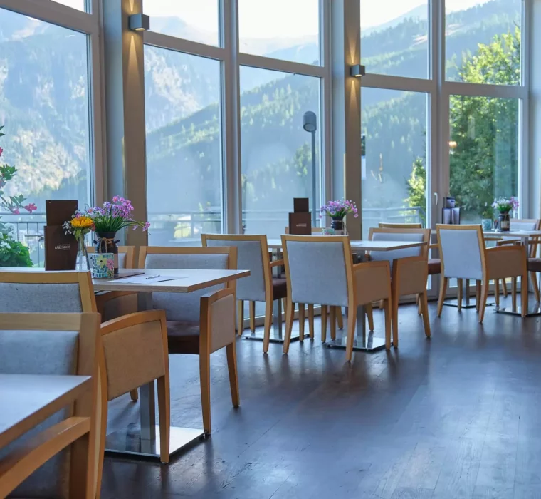 Helles Restaurant mit Holztischen und Stühlen, Blumen auf den Tischen, großen Fenstern und Blick auf Berge in Bad Gastein