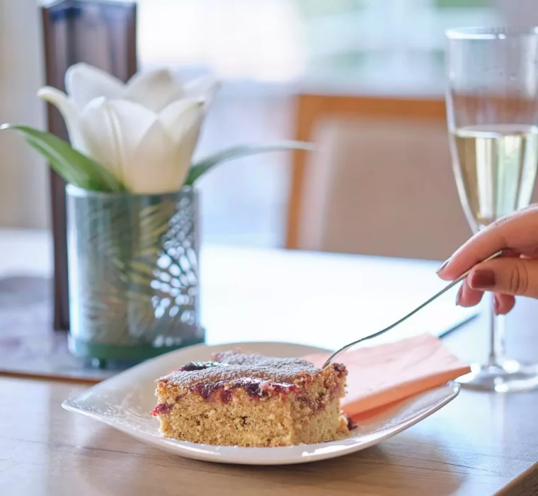 Stück Kuchen auf weißem Teller mit Gabel, Glas Sekt und weiße Blume auf Holztisch im Kurhotel Bärenhof Bad Gastein