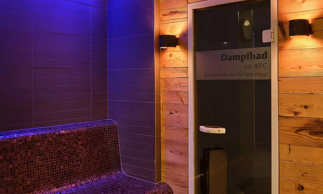 Innenansicht eines Dampfbads mit Mosaik-Sitzbank, Holzwand und beleuchteter Glastür im Gesundheitszentrum Bärenhof Bad