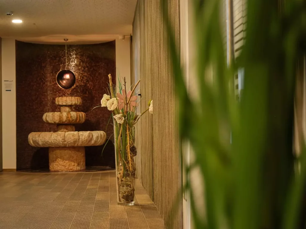 Flur im Gesundheitszentrum Bärenhof Bad Gastein mit dekorativem Steinbrunnen und Vase mit Blumen vor Vorhängen