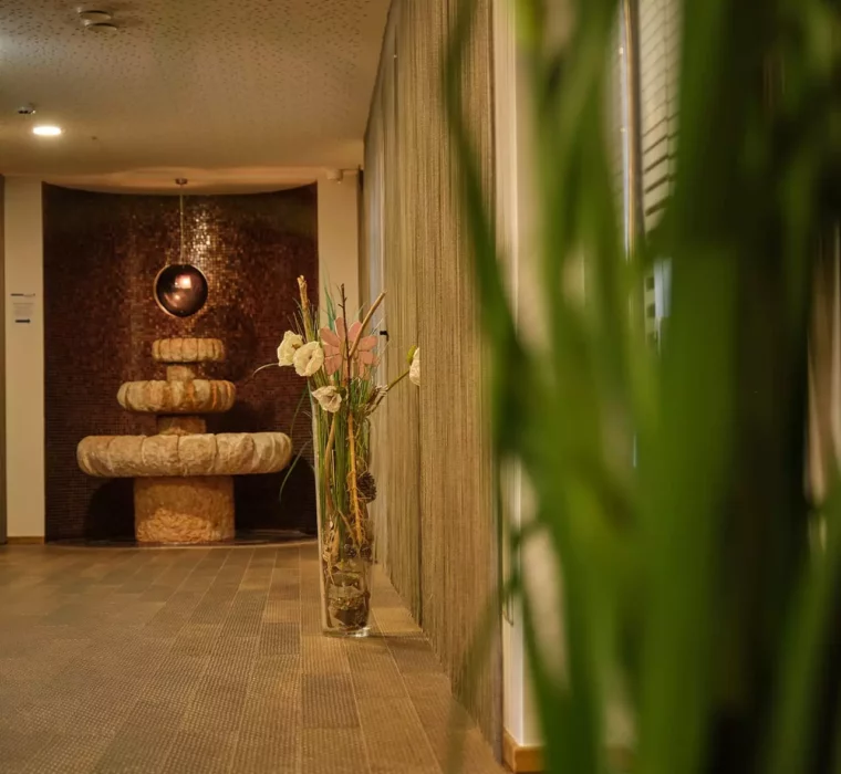 Flur im Gesundheitszentrum Bärenhof Bad Gastein mit dekorativem Steinbrunnen und Vase mit Blumen vor Vorhängen
