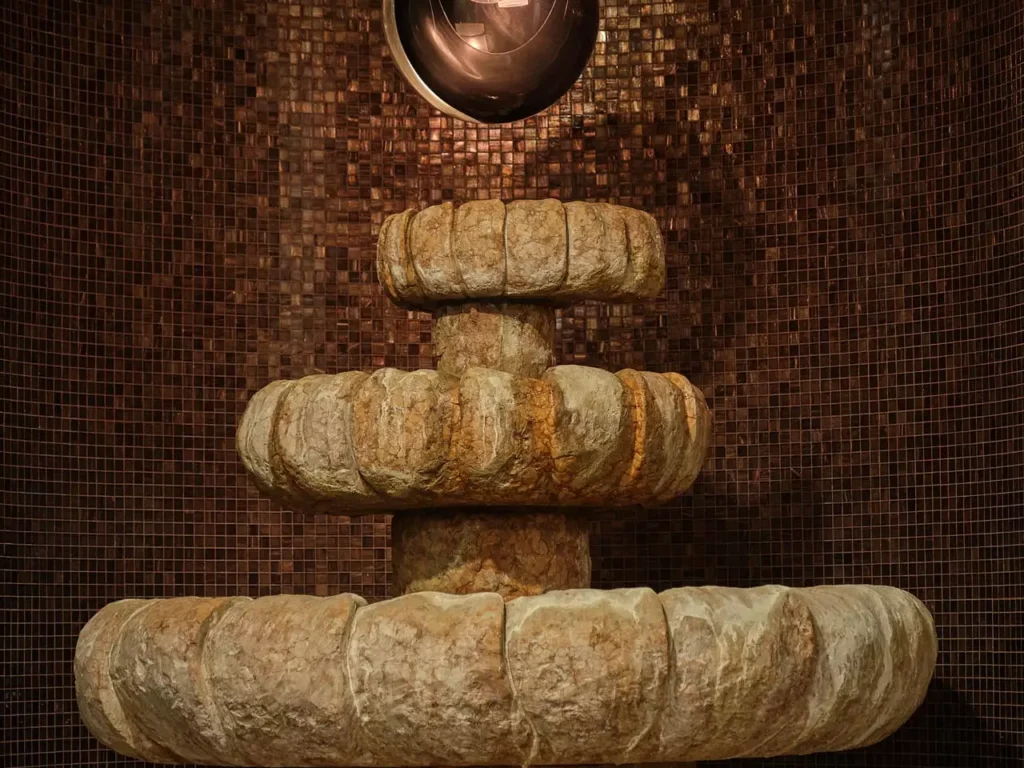 Steinerner dreistufiger Brunnen vor braun glänzender Mosaikwand im Gesundheitszentrum Bärenhof Bad Gastein