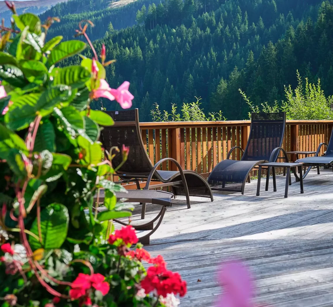 Sonnige Holzterrasse mit Liegestühlen und bunten Blumen vor bewaldeten Bergen im Gesundheitszentrum Bärenhof Bad Gastein