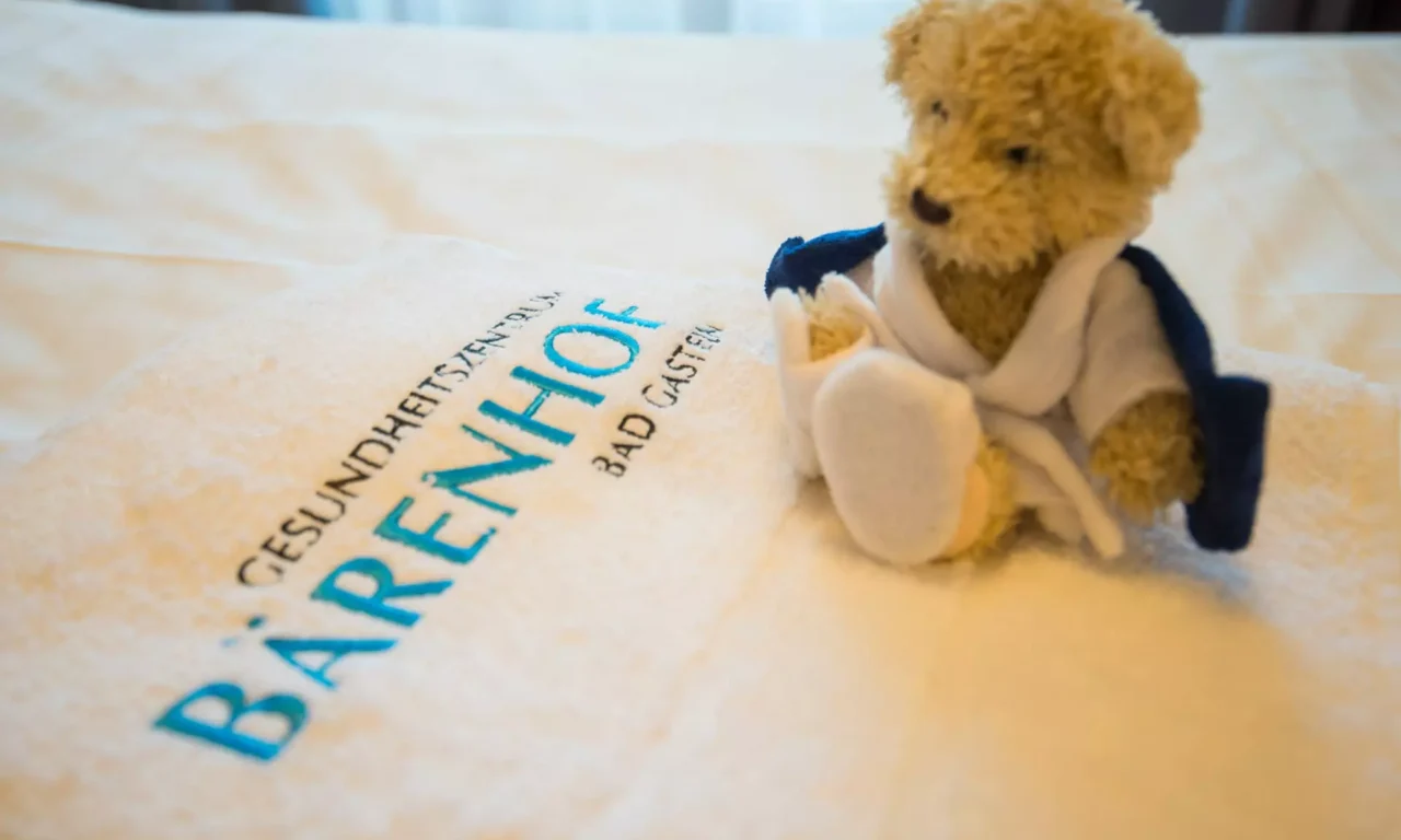 Kuscheliger Teddybär mit Bademantel sitzt auf einem weißen Handtuch mit Stickerei Gesundheitszentrum Bärenhof Bad Gastein