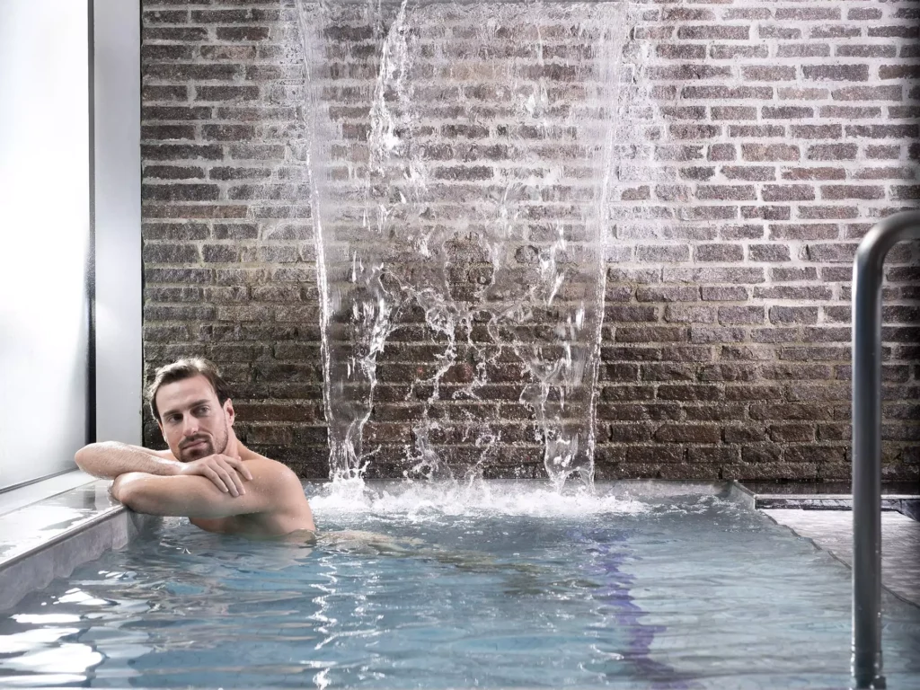 Mann entspannt im Innenpool des Gesundheitszentrums Bärenhof Bad Gastein unter einem Wasserfall vor Backsteinwand