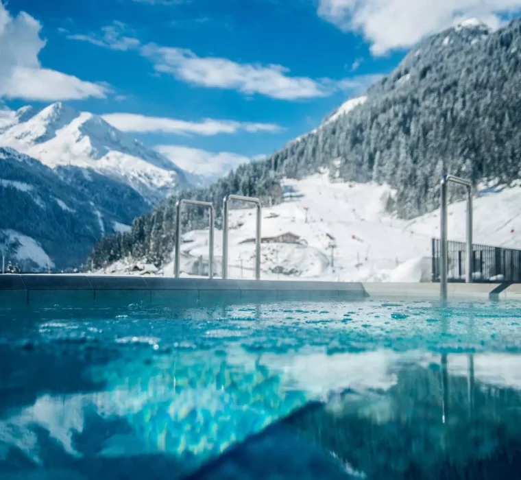 Außenpool mit klarem Wasser vor schneebedeckten Bergen und blauem Himmel im Gesundheitszentrum Bärenhof Bad Gastein