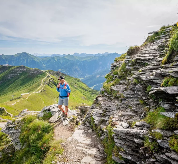 Wanderer mit rotem Rucksack auf Bergpfad neben steiniger Felswand mit Blick auf grüne Berge bei Bad Gastein