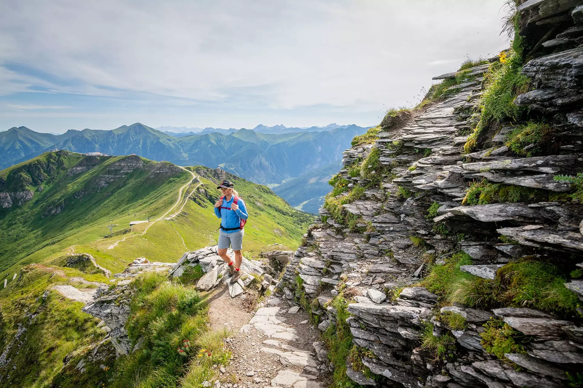 Wanderer mit rotem Rucksack auf Bergpfad neben steiniger Felswand mit Blick auf grüne Berge bei Bad Gastein
