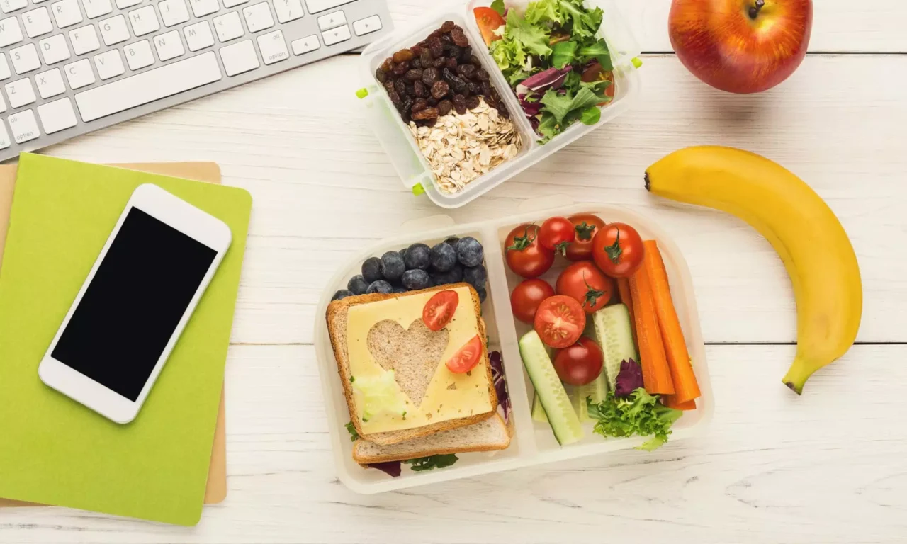 Gesunde Lunchbox mit Sandwich, Kirschtomaten, Gurken, Karotten, Blaubeeren, Salat, Apfel und Banane auf Schreibtisch mit