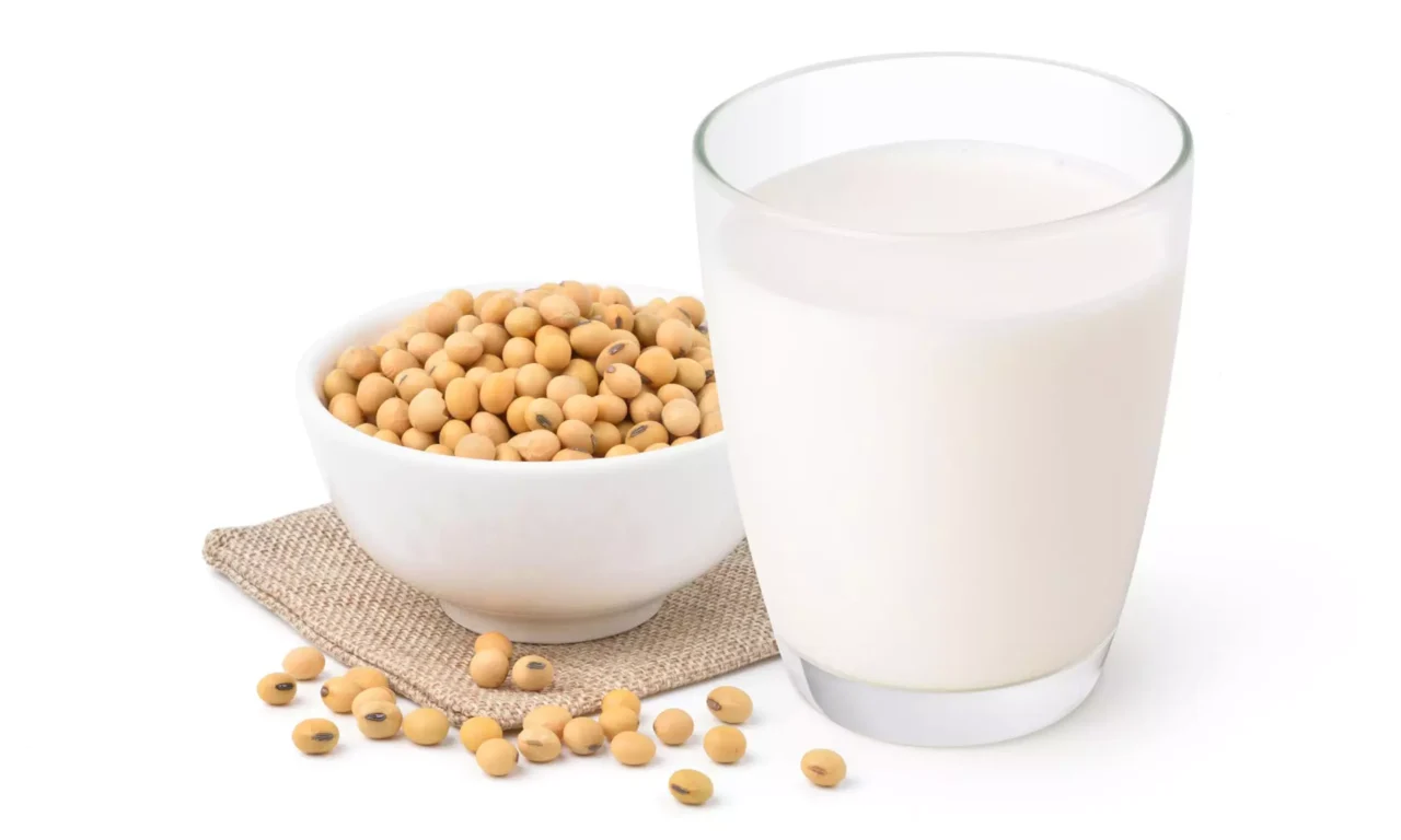 Glas mit Sojamilch neben weißer Schale mit Sojabohnen auf Stoffunterlage vor weißem Hintergrund