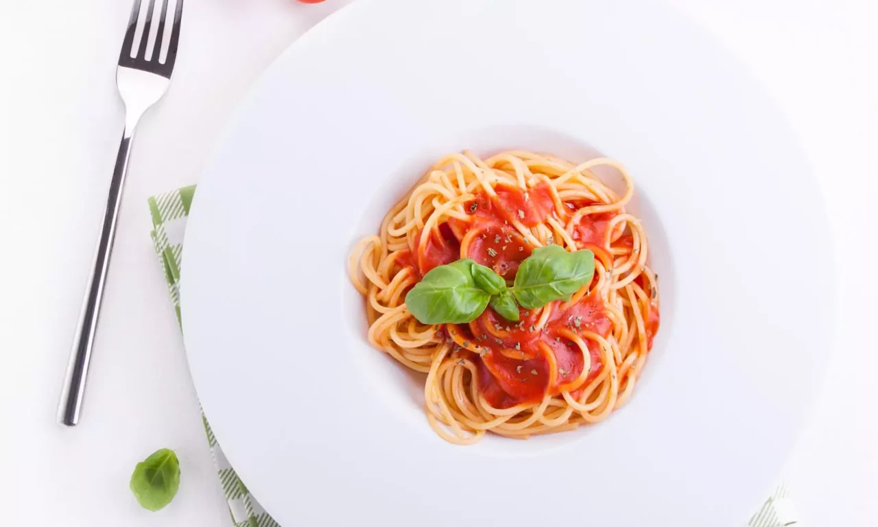 Spaghetti mit Tomatensauce und Basilikum auf weißem Teller, daneben Kirschtomaten, Knoblauch und Pfeffer im