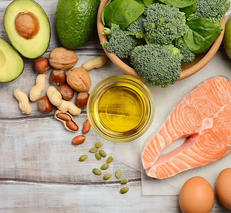 Gesunde Lebensmittel wie Avocado, Nüsse, Brokkoli, Lachs, Eier, grüner Apfel und Öl auf Holzuntergrund für Kuraufenthalt und