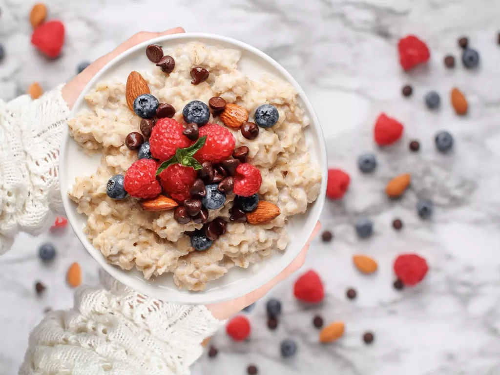 Schale Haferbrei mit Himbeeren, Blaubeeren, Mandeln und Schokotropfen, gehalten von Händen in weißem Strickpullover