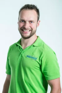 gesundheitszentrum-kurhotel-baerenhof-bad-gastein-therapien-2020-0045 Lächelnder Mann mit grünem Poloshirt mit Logo Gesundheitszentrum Bärenhof Bad Gastein vor hellem Hintergrund